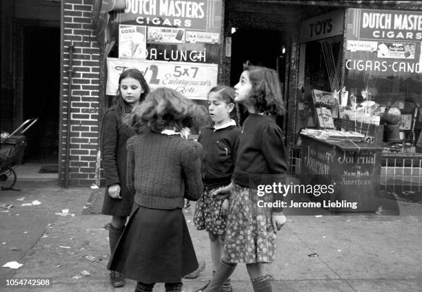 View of a group of girls on the sidewalk in front of Milrod Candy Store, Brooklyn, New York, New York, 1940. Signs advertise cigars, photo...