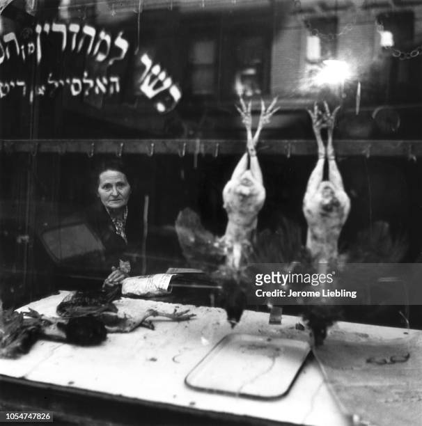 View of an unidentified elderly woman as she looks out through the window of a butcher's shop, where a pair of chickens hang upside, on Manhattan's...