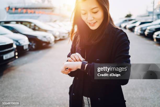 beautiful young asian woman checking time on smartwatch in city, in front of cars in outdoor carpark at sunset - asian and indian ethnicities smartwatch phone stock pictures, royalty-free photos & images