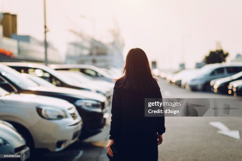 Rear view of young woman walking through outdoor car park in city looking for her car