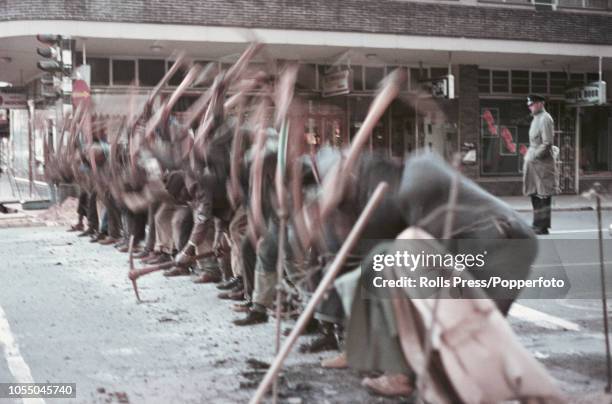 Watched by a white policeman, local black labourers use pick axes to break up the road surface of a city street in Johannesburg during the Apartheid...