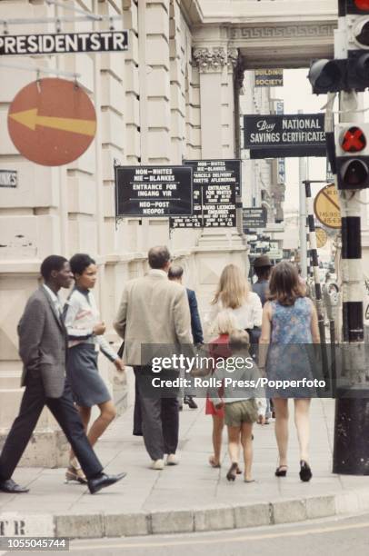 View of both black and white residents walking on a pavement past the entrance to a post office in the centre of Johannesburg during the Apartheid...