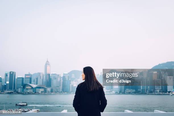 rear view of young woman standing against the promenade of victoria harbour overlooking the cityscape of hong kong - chinese person stock-fotos und bilder