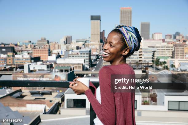 young woman smiling on rooftop while holding coffee cup - johannesburg foto e immagini stock