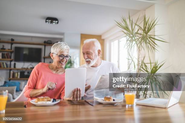 modern senior couple looking at newspaper and a tablet during breakfast - human settlement stock pictures, royalty-free photos & images