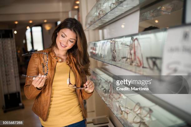 woman choosing glasses in optical store using mirror - buying eyeglasses stock pictures, royalty-free photos & images