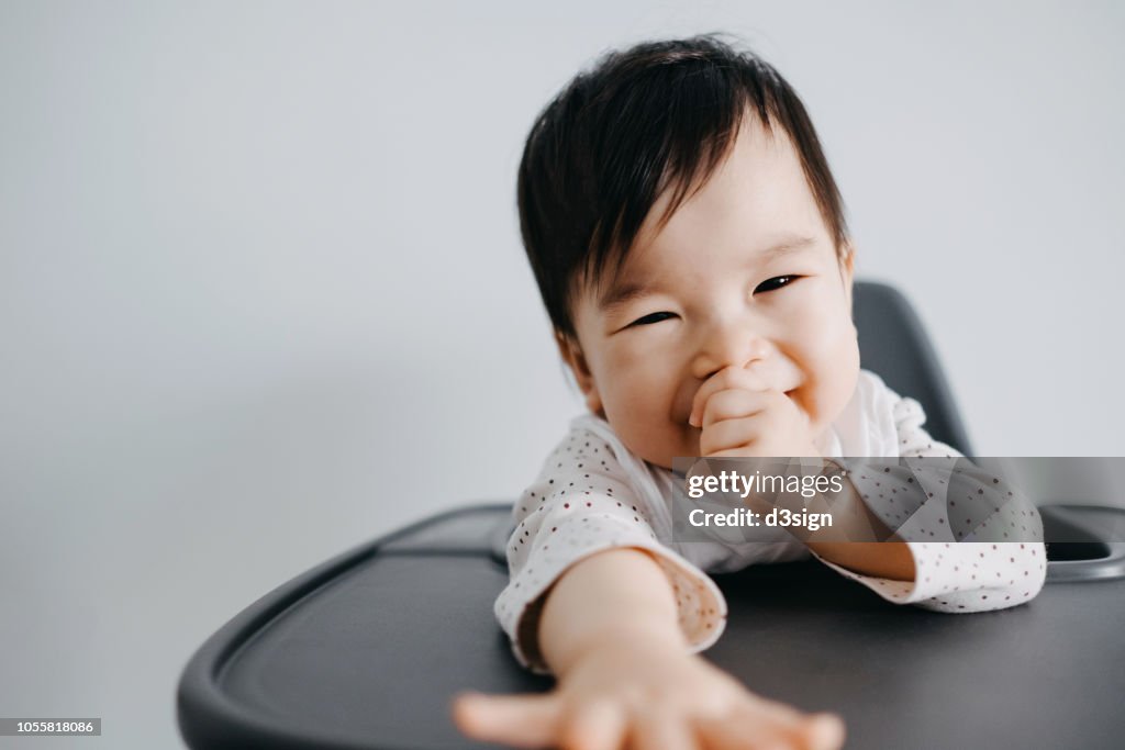 Cute baby girl sitting on high chair sucking her thumb and smiling joyfully