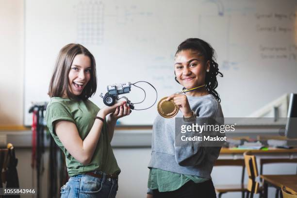 portrait of successful female teenage students holding robot and gold medal while standing in classroom at high school - teen awards bildbanksfoton och bilder