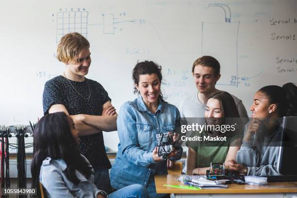 surprised teacher examining robot while sitting amidst students against whiteboard in classroom at high school - man robot stockfoto's en -beelden