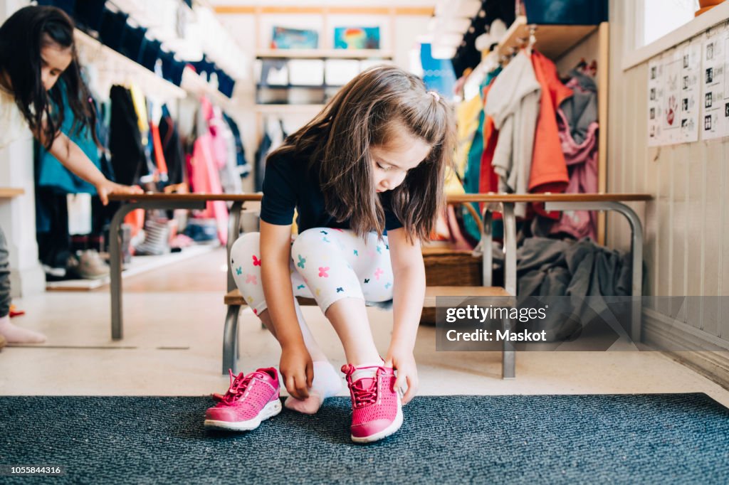 Full length of girl sitting on bench wearing shoes in cloakroom at preschool