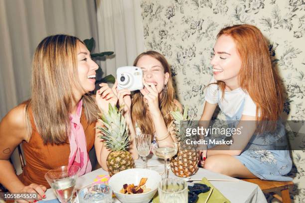 cheerful young woman holding camera while sitting amidst female friends at home during dinner party - photographing bildbanksfoton och bilder