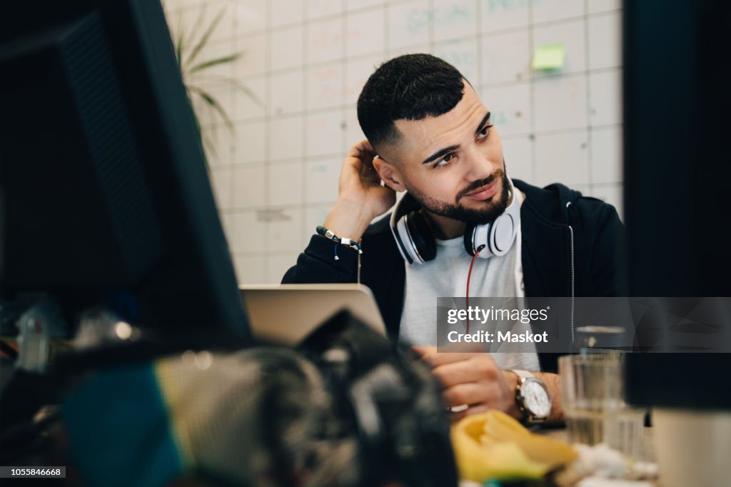 Confused young male computer programmer scratching head while sitting with laptop at creative office