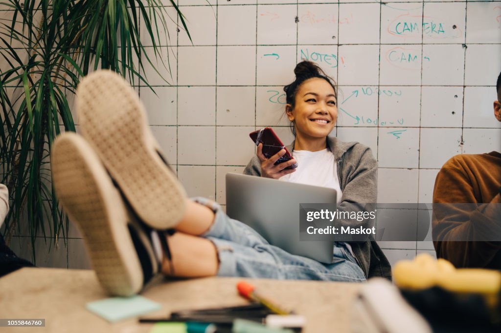 Smiling young businesswoman sitting with feet up on desk using wireless technologies by colleague against wall at office