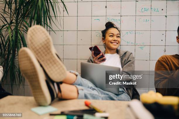 smiling young businesswoman sitting with feet up on desk using wireless technologies by colleague against wall at office - soles pose stock pictures, royalty-free photos & images