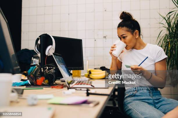 young businesswoman drinking coffee while sitting at desk in small creative office - design professional stockfoto's en -beelden
