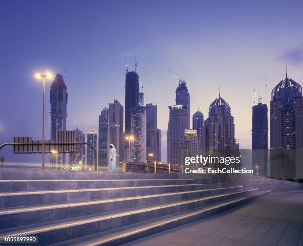 arab man in traditional robe waiting in front of dubai skyline at sunset - modern tradition stock pictures, royalty-free photos & images