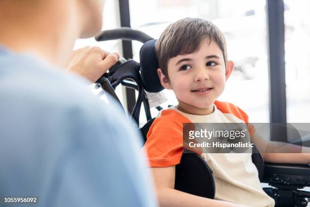 schöner kleiner junge am rollstuhl lächelnd und mit bewunderung auf seine unrecongizable-physiotherapeut - child in wheelchair stock-fotos und bilder