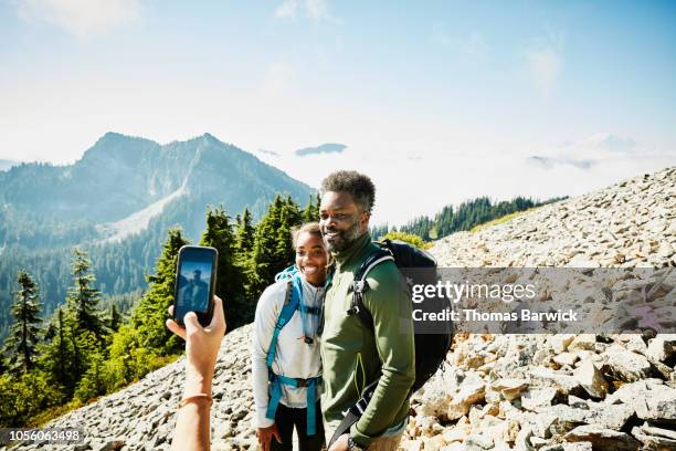 woman taking photo of father and daughter with smart phone during hike in mountains - 3 teenagers mobile outdoors stock-fotos und bilder