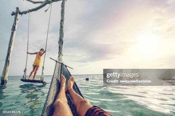 young couple swinging on the beach by the sea, beautiful and idyllic landscape. people travel romance vacations concept. personal perspective of man on sea hammock and girlfriend on sea swing. - couple swinging stock pictures, royalty-free photos & images