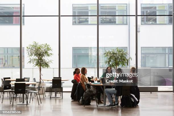 full length of students talking while sitting at table in university - campus - fotografias e filmes do acervo