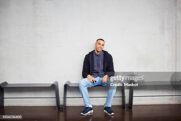 full length portrait of smiling young student sitting on bench against wall in university - guy with phone full image ストックフォトと画像