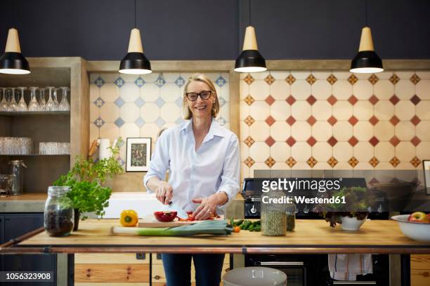 portrait of smiling mature woman chopping bell pepper in kitchen - kitchen bench top stock pictures, royalty-free photos & images