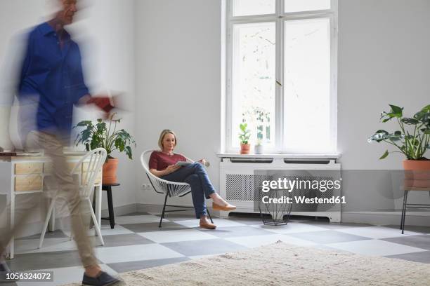 mature woman reading magazine at home with man passing by - blurred motion home stock pictures, royalty-free photos & images