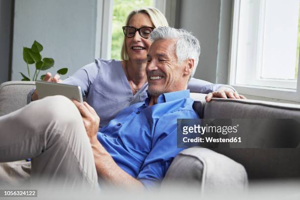 happy mature couple sitting on couch at home sharing tablet - 50 59 years stock pictures, royalty-free photos & images