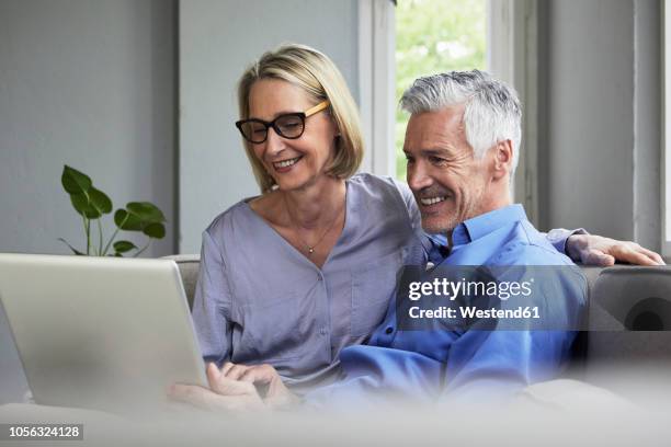 happy mature couple sitting on couch at home sharing laptop - couple 50 55 laptop stock-fotos und bilder