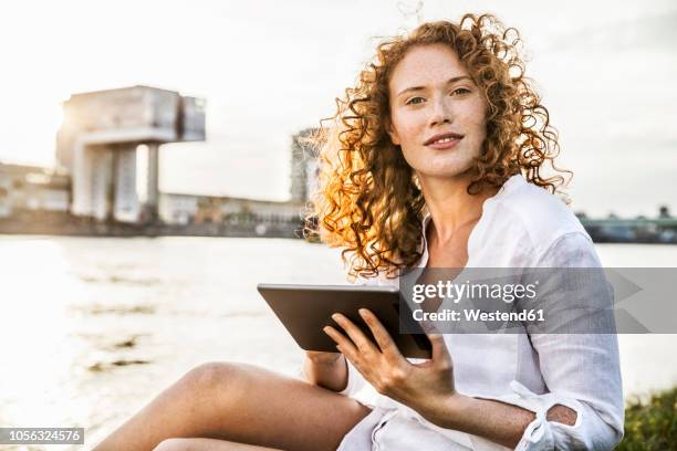 germany, cologne, portrait of young woman with tablet sitting at riverside - anelzinho - fotografias e filmes do acervo