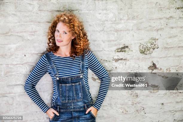 portrait of smiling young woman wearing denim dungarees leaning against white brick wall - jeans latzhose frau stock-fotos und bilder