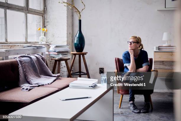 pensive woman sitting on arm chair in loft looking through window - woman thinking hand on chin stock pictures, royalty-free photos & images
