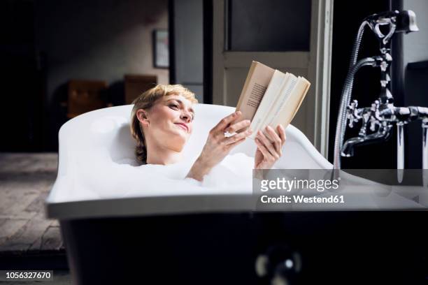 portrait of relaxed woman taking bubble bath in a loft reading a book - low key stock-fotos und bilder