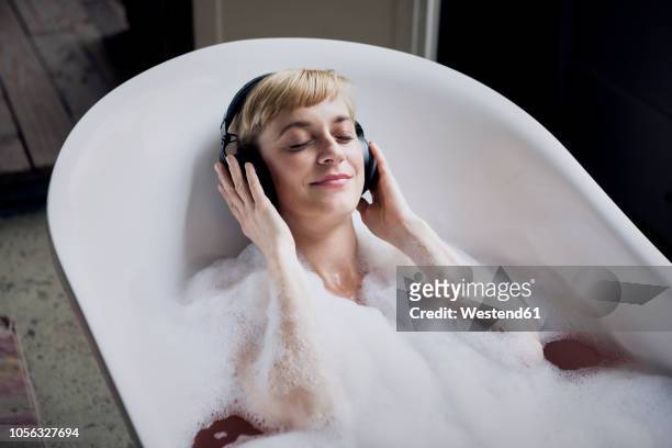 blond woman taking bubble bath in a loft listenung music with headphones - schaumbad stock-fotos und bilder