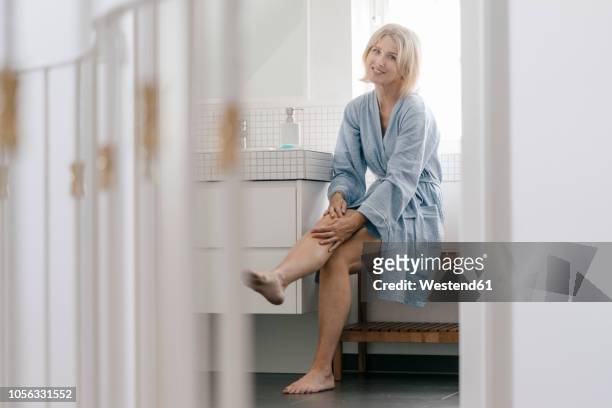 portrait of smiling mature woman sitting in bathroom touching her legs - older woman legs fotografías e imágenes de stock