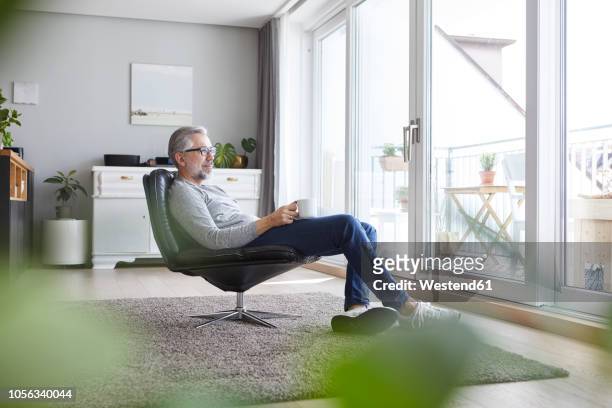 mature man sitting on leather chair in his living room relaxing with cup of coffee - man side view stock pictures, royalty-free photos & images