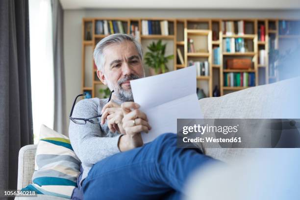 mature man sitting on couch at home reading letter - reading document stock pictures, royalty-free photos & images