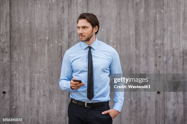 businessman standing at concrete wall holding smartphone - om zich heen kijken stockfoto's en -beelden
