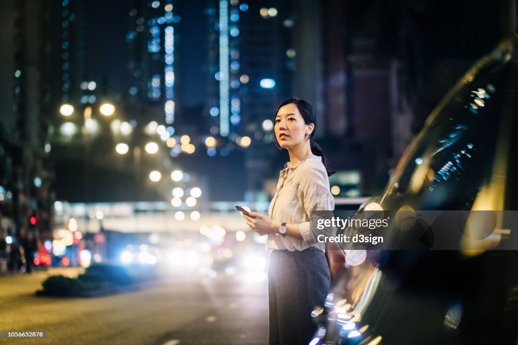 Young Asian woman using mobile app on smart watch to arrange taxi ride in downtown city street, with blurry traffic scene as background