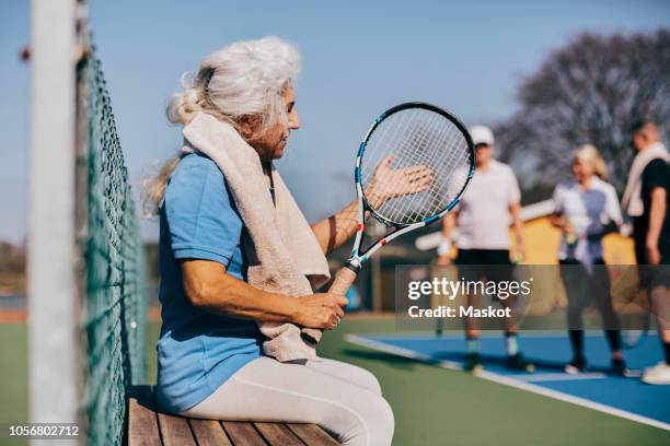 senior woman holding tennis racket while sitting on bench at tennis court - senior tennis stock pictures, royalty-free photos & images