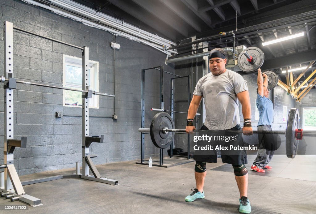 Two Latino athletes, the young man on the front, and the senior 55 years old man in the backstage, lifting heavy weights in the gym