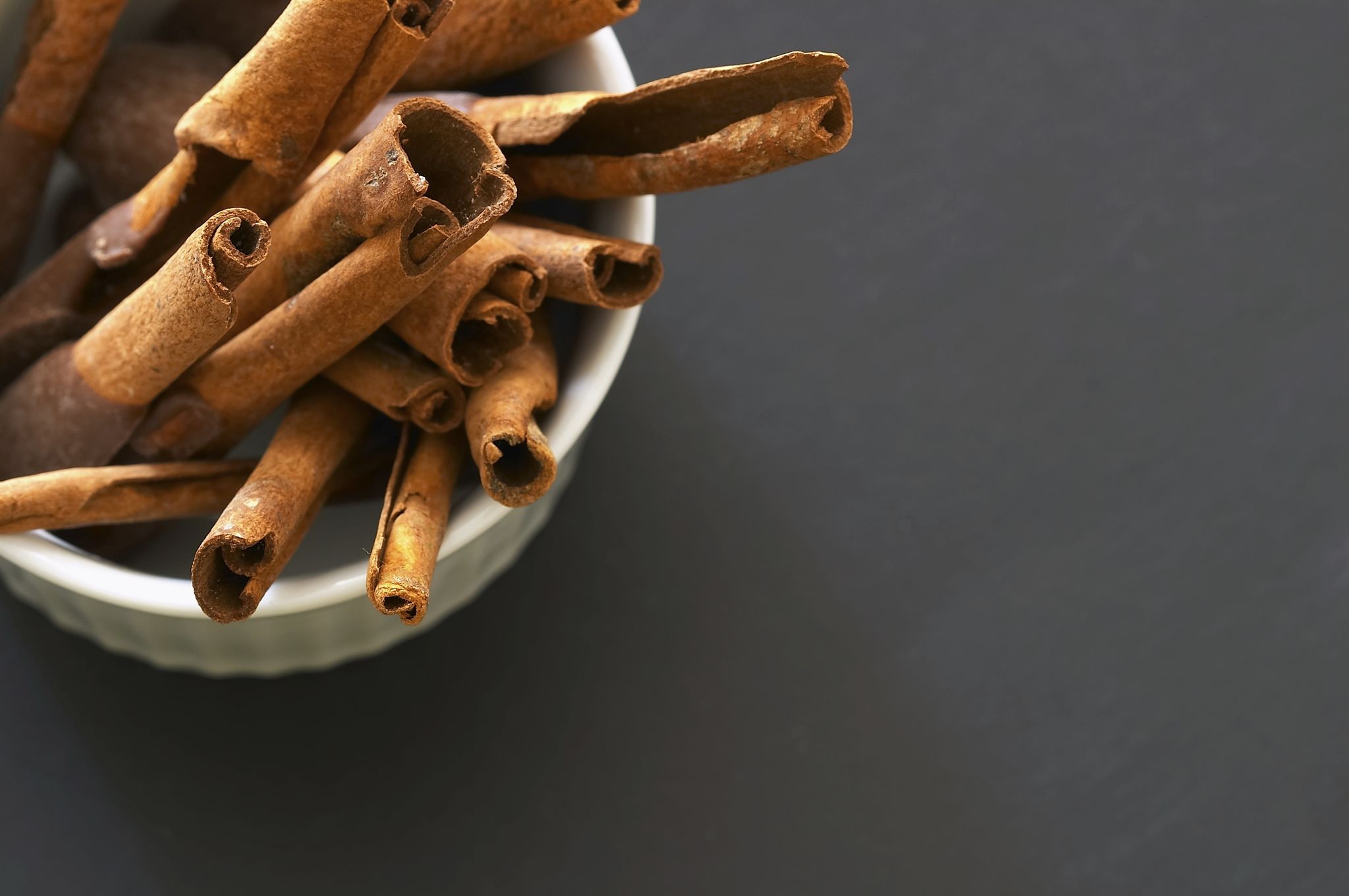 A white bowl filled with dried cinnamon sticks, placed on a dark, textured surface. A white bowl filled with dried cinnamon sticks, placed on a dark, textured surface.