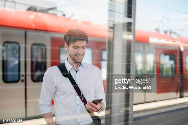 smiling businessman on station platform with earphones and cell phone - passagierzug stock-fotos und bilder