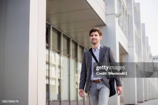 smiling businessman with crossbody bag in the city on the move - grauer anzug stock-fotos und bilder