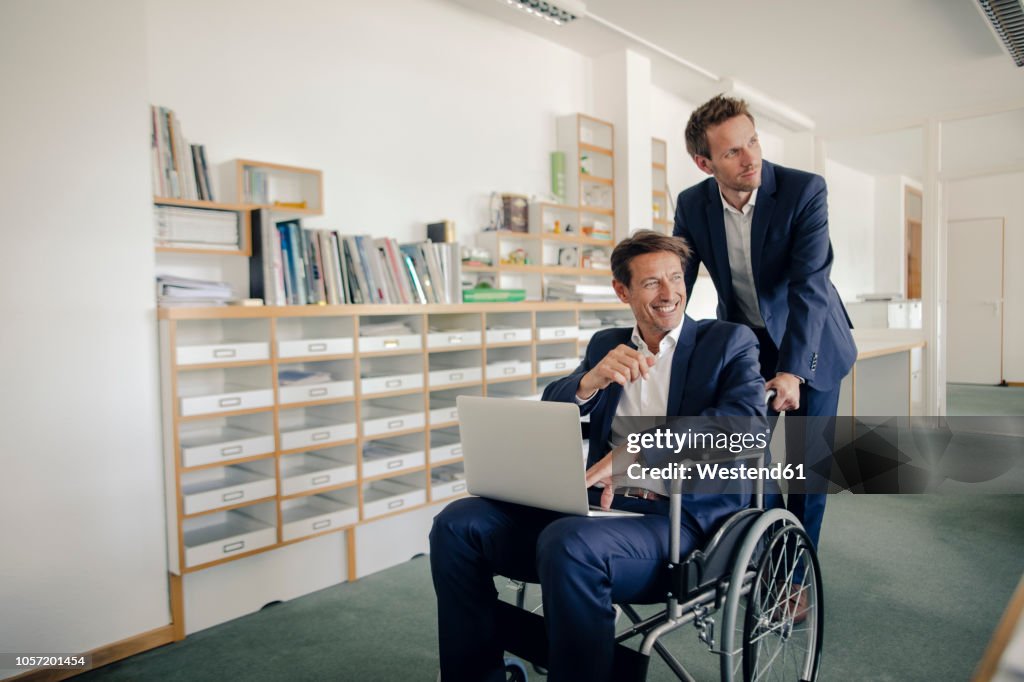 Colleague pushing businessman, sitting in wheelchai¥r