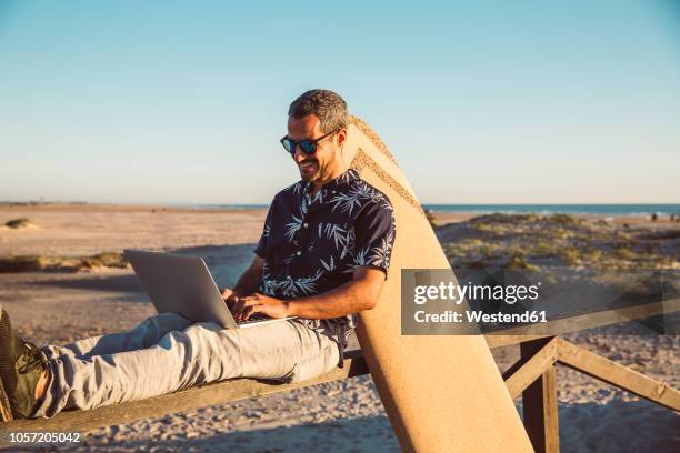 man sitting at the beach, using laptop, with surfboard leaning on fence - sitting on surfboard stockfoto's en -beelden