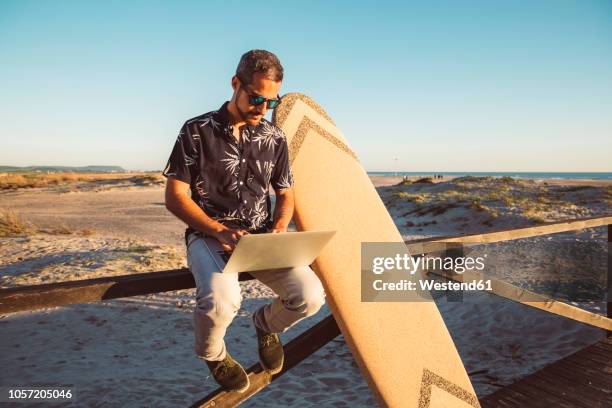 man sitting at the beach, using laptop, with surfboard leaning on fence - surfing the net fotografías e imágenes de stock