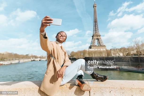 france, paris, woman sitting on bridge over the river seine with the eiffel tower in the background taking a selfie - turismo foto e immagini stock