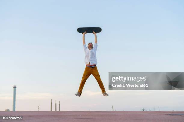young man jumping in the air holding a skateboard - hochhalten stock-fotos und bilder