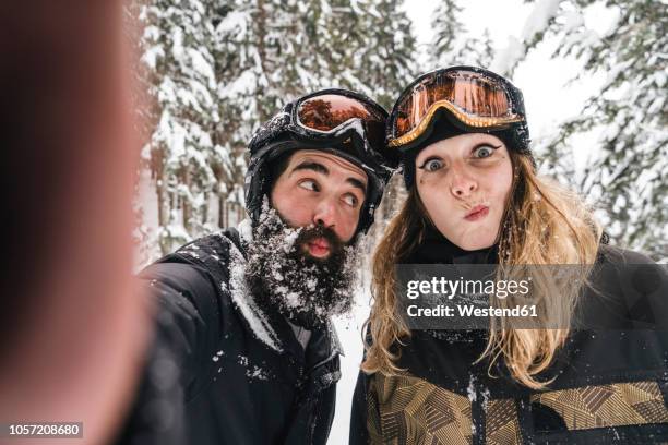 selfie of happy couple in skiwear grimacing in winter forest - female puckered lips stockfoto's en -beelden